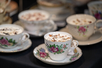 A table with multiple ceramic traditional Victorian white teacups with various rose and flora patterns on the porcelain beverage cups for sale. The wax in the cups has scented dried flower petals.