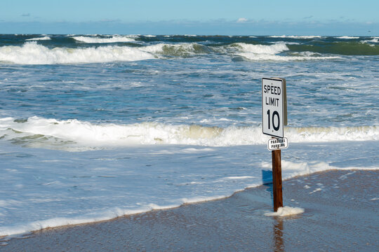 A speed limit of 10 sign and aparallel parking sign on a wooden post on a sandy beach. There's a stormy beach with waves and currents rolling on shore. The warning signs are funny. The beach is empty.
