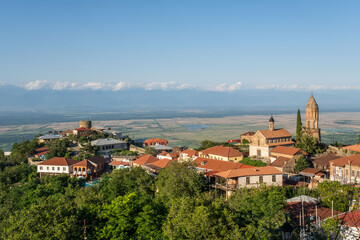 Obraz premium A charming European town with red-roofed buildings, a church with a tall steeple, rolling hills, and majestic mountains in the background