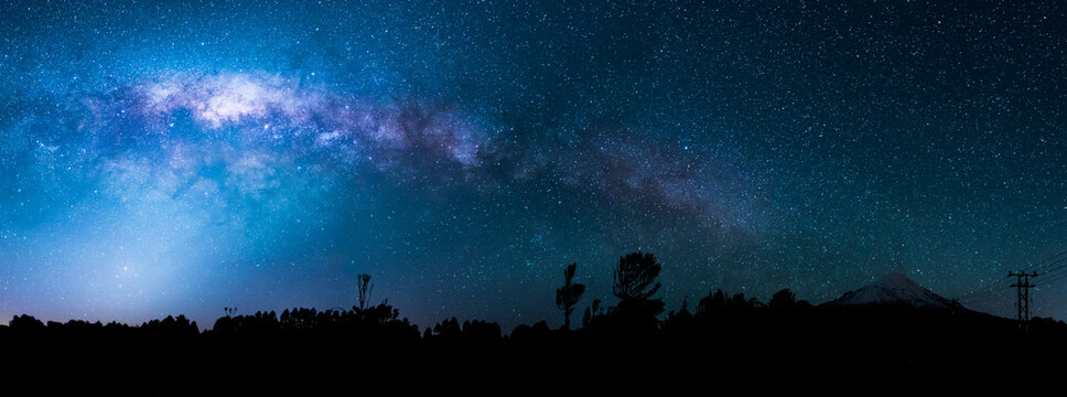 Wide panorama of the night sky showing the Milky Way and the Galactic Centre. In the foreground are various trees in silhouette and, at left, Mount Taranaki (Egmont).