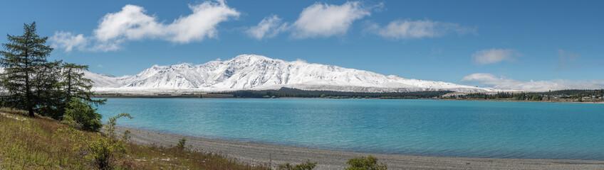 Obraz premium Wide panorama of the turquoise waters of Lake Tekapo, New Zealand. Snow-capped mountains in the background and gravel beach in the foreground.