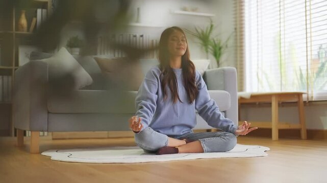 Happy young Asian woman practicing yoga and meditation at home sitting on floor in living room in lotus position and relaxing with closed eyes. Mindful meditation and wellbeing concept