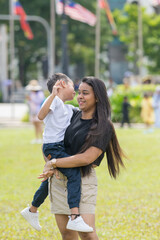 A thirty-something Indian Malaysian mother joyfully playing on the expansive lawn at Merdeka Square in Kuala Lumpur, Malaysia, while holding her seven-year-old son.