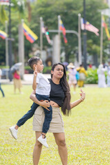 Naklejka premium A thirty-something Indian Malaysian mother joyfully playing on the expansive lawn at Merdeka Square in Kuala Lumpur, Malaysia, while holding her seven-year-old son.