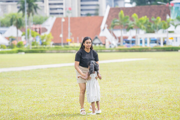 A thirty-something Indian Malaysian mother joyfully playing on the expansive lawn at Merdeka Square in Kuala Lumpur, Malaysia, while holding and running around with her five-year-old daughter.