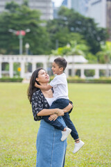 A 30-something Indian Malaysian mother joyfully playing on the expansive lawn at Merdeka Square in Kuala Lumpur, Malaysia, while holding her 7-year-old son.