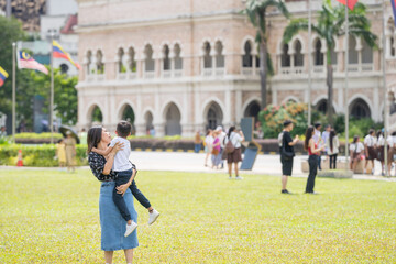 A 30-something Indian Malaysian mother joyfully playing on the expansive lawn at Merdeka Square in Kuala Lumpur, Malaysia, while holding her 7-year-old son.