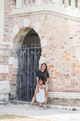 A 30-something Indian Malaysian mother and her 7-year-old son and 5-year-old daughter spending time joyfully in front of Western-style brick historical building in Kuala Lumpur, Malaysia, on hot day.
