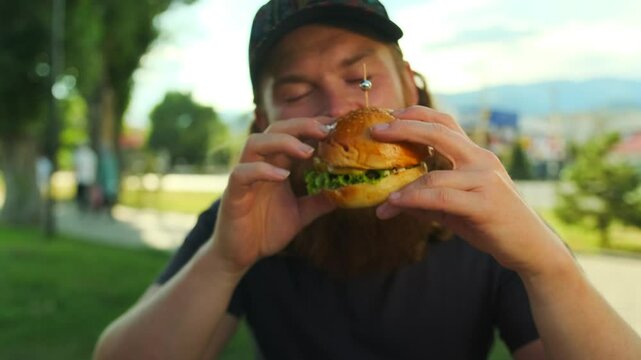 Gluttonous bearded young man eating a juicy burger with meat enjoying the taste, while sitting in a fast food cafe on the street.