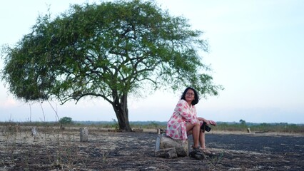 A woman sitting on a trunk in a savanna with a big tree on the background