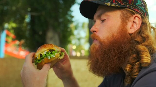 Bearded hungry man eating an appetizing burger sitting in the cafe outdoors on the blurred background. Slow motion.