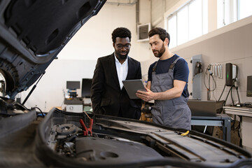 Mechanic in overalls discussing car maintenance with businessman using tablet. Scene in modern auto...