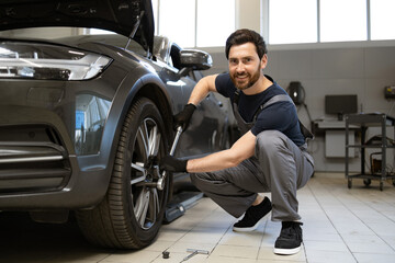 Fototapeta premium Mechanic in auto repair shop working on car wheel, using wrench. Professional mechanic delivering car maintenance service with smile. Modern workshop environment with car repair tools.