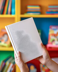 A person is holding a white book in their hands. The book is placed on a shelf with other books. The scene suggests a library or a reading area where people can find books to read