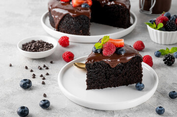 Delicious chocolate cake with chocolate glaze and fresh berries on a white plate on a gray concrete background.