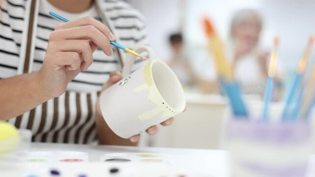 Closeup of hands of talented female artisan creating own unique ceramic items in pottery workshop, painting mug with engobe and brush. Craft activity concept
