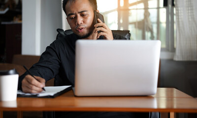 Handsome young Asia man in formal wear talking on smartphone, enjoying conversation at workplace. Young businessman using laptop in his office. 
