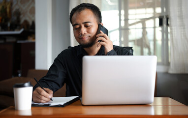 Handsome young Asia man in formal wear talking on smartphone, enjoying conversation at workplace. Young businessman using laptop in his office. 
