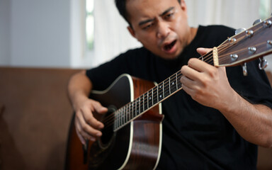 Asian man with  sitting on a sofa in his home's living room, picking up a guitar.