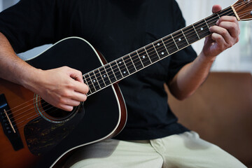 Fototapeta premium Asian man with sitting on a sofa in his home's living room, picking up a guitar.