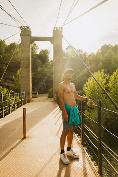 Shirtless sporty guy standing on a bridge over a lake during sunset