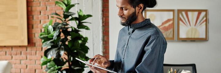Website header shot of young Indian man searching information via huge tablet in cozy office