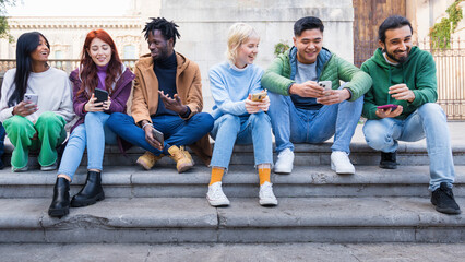 Fototapeta premium Diverse group of young friends using smartphones outdoors, multicultural millennials socializing, digital connectivity, urban lifestyle, inclusive friendship