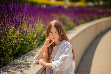A young girl is basking in the warm rays of the sun, enjoying the beauty of the park, overgrown with bright purple sage. Her white shirt and blue trousers complete the landscape