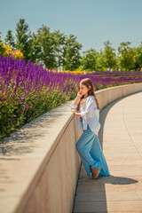 A young girl is basking in the warm rays of the sun, enjoying the beauty of the park, overgrown with bright purple sage. Her white shirt and blue trousers complete the landscape