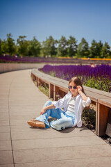 A young girl is basking in the warm rays of the sun, enjoying the beauty of the park, overgrown with bright purple sage. Her white shirt and blue trousers complete the landscape