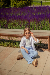 A young girl is basking in the warm rays of the sun, enjoying the beauty of the park, overgrown with bright purple sage. Her white shirt and blue trousers complete the landscape