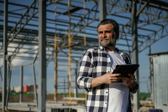 Wireless device, holding digital tablet in hands. Senior worker is near the unfinished greenhouse, working