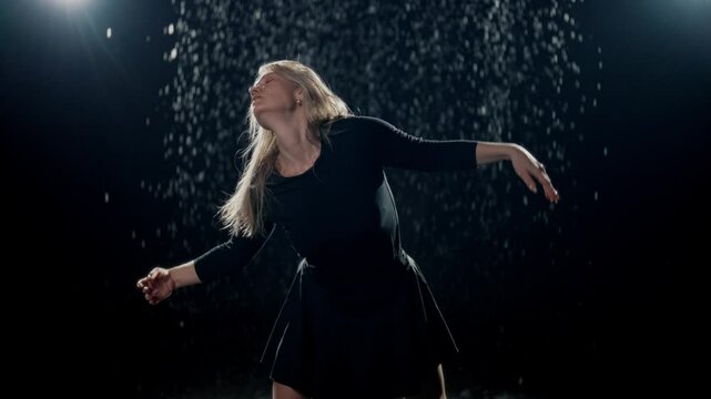 Excited woman dancer moving passionately in dark studio with false rain or snow. Blonde lady dancing in rain in night, beautiful female dancer performing modern choreography, graceful female body