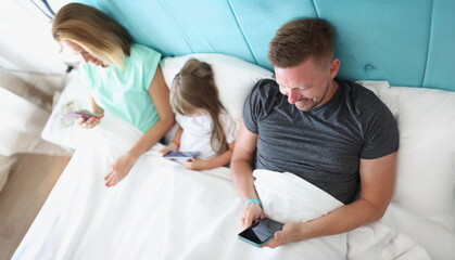 Obraz premium Top view of young family with kid using gadgets while lying together in bed. Leisure and fun time, happy family, parenthood and childhood