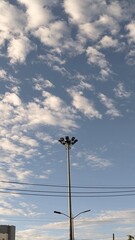 A tall light pole stands against a blue sky dotted with scattered clouds. Below, power lines stretch across, framing the scene with a touch of urban simplicity.