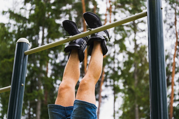 A mans legs in gravity boots hangs on a horizontal bar outdoors on a summer day.