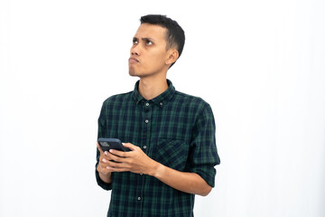 Asian man wearing a green checkered casual shirt is thinking while holding a smartphone. Isolated white background.