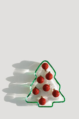 Green colored glass plate form as New Year tree with sparkling red Christmas balls instead of food, sunlight, shadow, star glare on white background. Winter holiday dinner concept, minimal