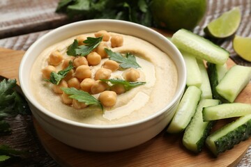 Tasty hummus with chickpeas in bowl and cucumbers on table, closeup