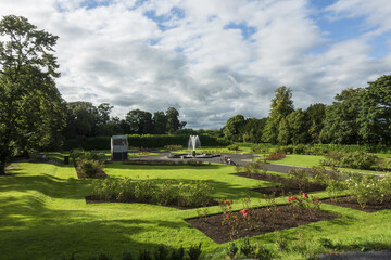 Summer and afternoon view of trees and rose flowers on the garden with tourists on bench besides fountain at Kilkenny Castle near Kilkenny, Ireland
