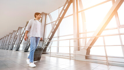 Enjoying vacation. Cheerful man walking with luggage in airport terminal, looking at sunset in window, empty space