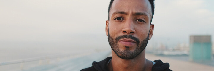 Obraz premium Close-up, young bearded male athlete standing on the embankment, looking at the sea. Guy turns his head and looks at the camera, panorama