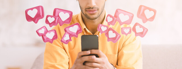 A man sits on a couch, holding a smartphone in his hands, looking down at the screen. A number of...