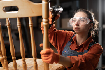 Young professional carpenter woman with an electric drill fixing wooden chair in workshop.