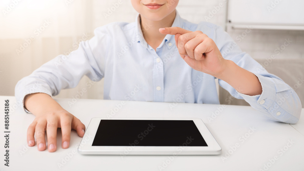 Poster A young boy sits at a white desk in a bright room. He is wearing a light blue button-down shirt and is using a digital tablet with a black screen. He is pointing with his index finger at the tablet.