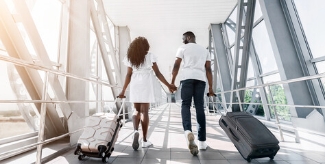 Black couple walks hand-in-hand through an airport terminal, each pulling a suitcase behind them....