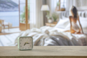 Alarm clock on wooden table top. Blurred background of a woman getting up in fresh early morning.   Table with a blurred bedroom background. Copy space for products.
