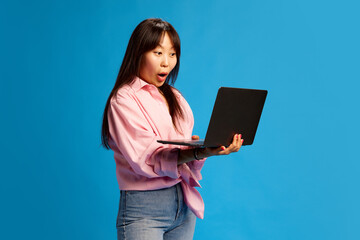 Young Korean woman in pink shirt looking on laptop with shocked positive face and excitement against blue studio background. Good news. Concept of human emotions, youth, lifestyle, business, sales