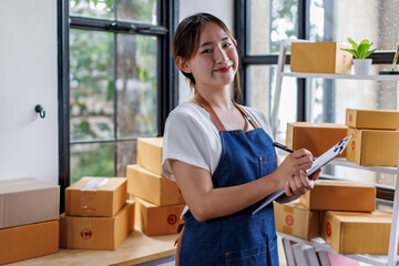 Startup SME small business entrepreneur of freelance Asian woman wearing apron using laptop and box to receive and review orders online to prepare to pack sell to customers, online sme business ideas.