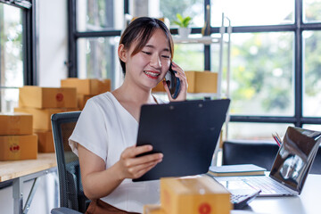 Young asia seller woman happy work on laptop busy cellphone call chat to customer at home office desk use digital tablet computer in SME online store social e-commerce sale page service web support.
